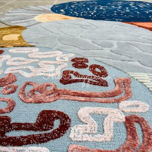 May include: A round, abstract rug with a light blue background and various shapes in shades of brown, white, and orange. The rug features a textured design and the text "the ruggesso" in the upper right corner.
