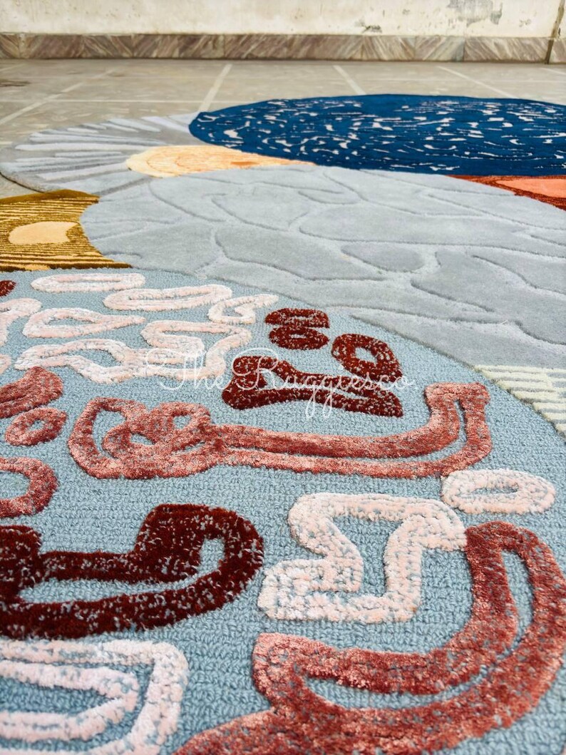 May include: A round, abstract rug with a light blue background and various shapes in shades of brown, white, and orange. The rug features a textured design and the text "the ruggesso" in the upper right corner.