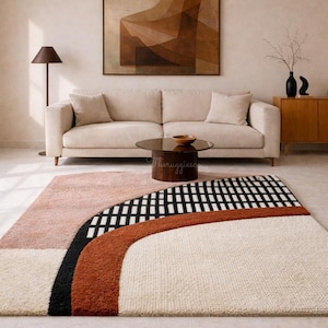 Op de afbeelding: Een modern vloerkleed met een geometrisch ontwerp. Het vloerkleed heeft een crèmekleurige basis met secties van roze, zwarte en roestkleurige gebogen lijnen en een zwart-wit geblokt patroon. Het vloerkleed staat in een woonkamer.