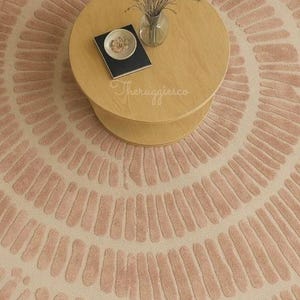 Pu&ograve; includere: Un tappeto rotondo con sfondo crema e un motivo di linee rosa curve che si irradiano dal centro. Un tavolino da caff&egrave; in legno chiaro &egrave; posto sul tappeto, con un vaso di steli secchi, un piattino e un libro.