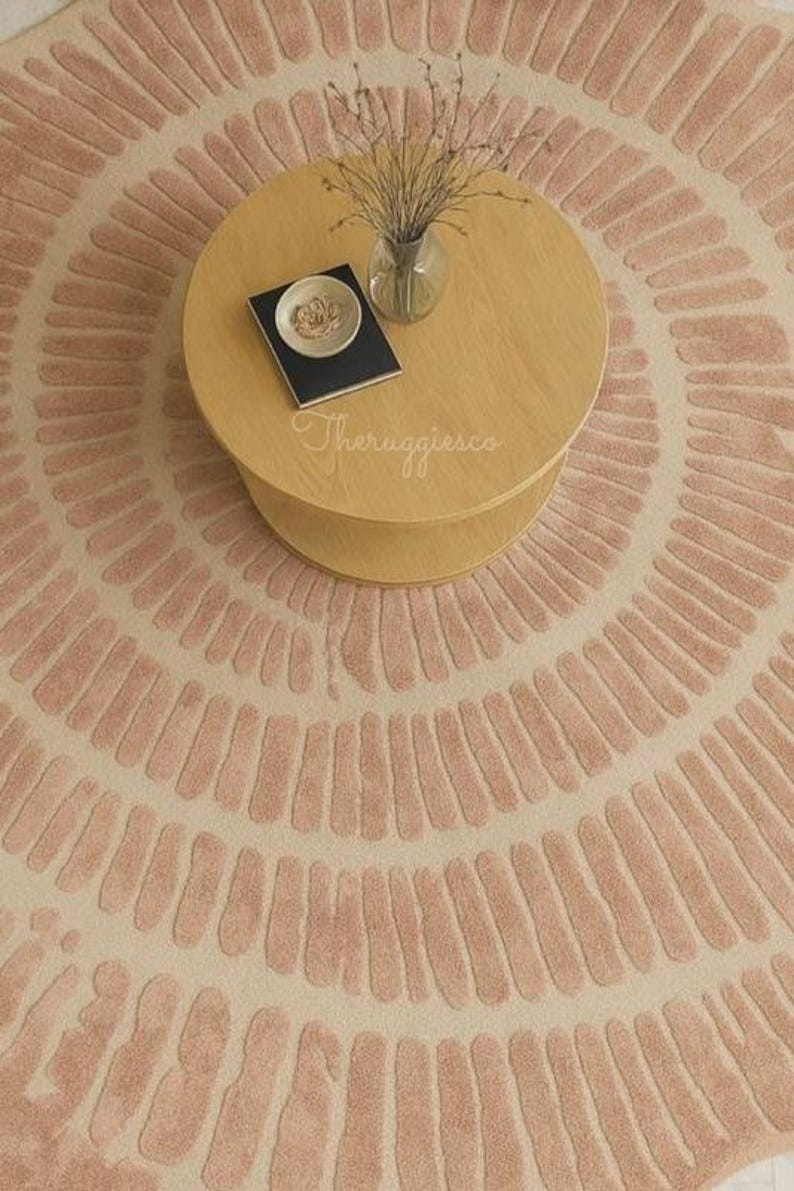 Pu&ograve; includere: Un tappeto rotondo con sfondo crema e un motivo di linee rosa curve che si irradiano dal centro. Un tavolino da caff&egrave; in legno chiaro &egrave; posto sul tappeto, con un vaso di steli secchi, un piattino e un libro.