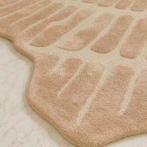 Pu&ograve; includere: Primo piano di un tappeto beige dal design unico. Il tappeto presenta un motivo di forme allungate e strutturate in un colore beige pi&ugrave; chiaro su uno sfondo beige pi&ugrave; scuro. Il tappeto ha un aspetto morbido e felpato e un bordo arrotondato.