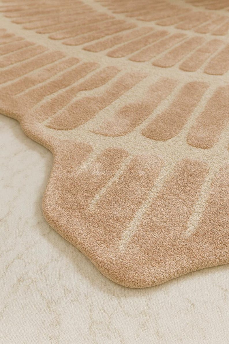 Pu&ograve; includere: Primo piano di un tappeto beige dal design unico. Il tappeto presenta un motivo di forme allungate e strutturate in un colore beige pi&ugrave; chiaro su uno sfondo beige pi&ugrave; scuro. Il tappeto ha un aspetto morbido e felpato e un bordo arrotondato.