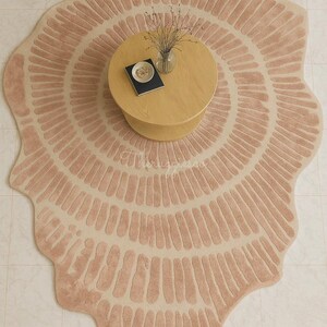 Pu&ograve; includere: Un grande tappeto rotondo con un design unico. Il tappeto presenta una combinazione di colori rosa chiaro e beige con un motivo radiale di linee verticali. Sono visibili anche un tavolino in legno e un divano beige.