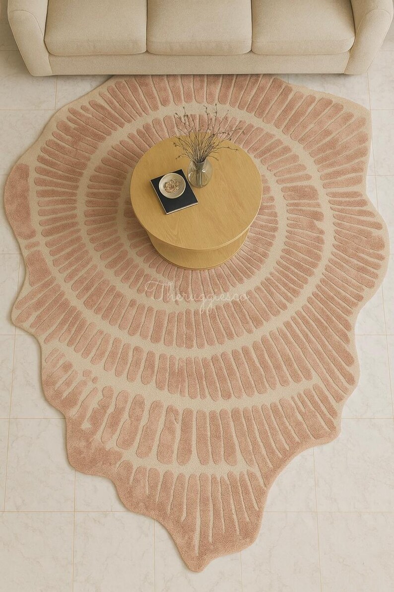 Pu&ograve; includere: Un grande tappeto rotondo con un design unico. Il tappeto presenta una combinazione di colori rosa chiaro e beige con un motivo radiale di linee verticali. Sono visibili anche un tavolino in legno e un divano beige.