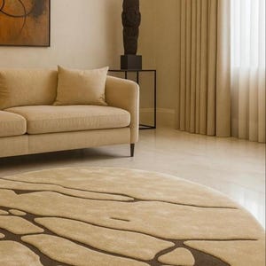 Pode incluir: Um tapete redondo bege com um padr&atilde;o abstrato castanho escuro domina o primeiro plano. Um sof&aacute; bege e almofadas s&atilde;o vis&iacute;veis, juntamente com uma escultura escura sobre uma mesa preta. A sala tem paredes e cortinas bege.