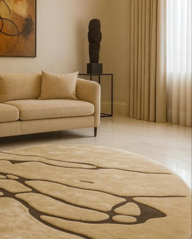 Pode incluir: Um tapete redondo bege com um padr&atilde;o abstrato castanho escuro domina o primeiro plano. Um sof&aacute; bege e almofadas s&atilde;o vis&iacute;veis, juntamente com uma escultura escura sobre uma mesa preta. A sala tem paredes e cortinas bege.