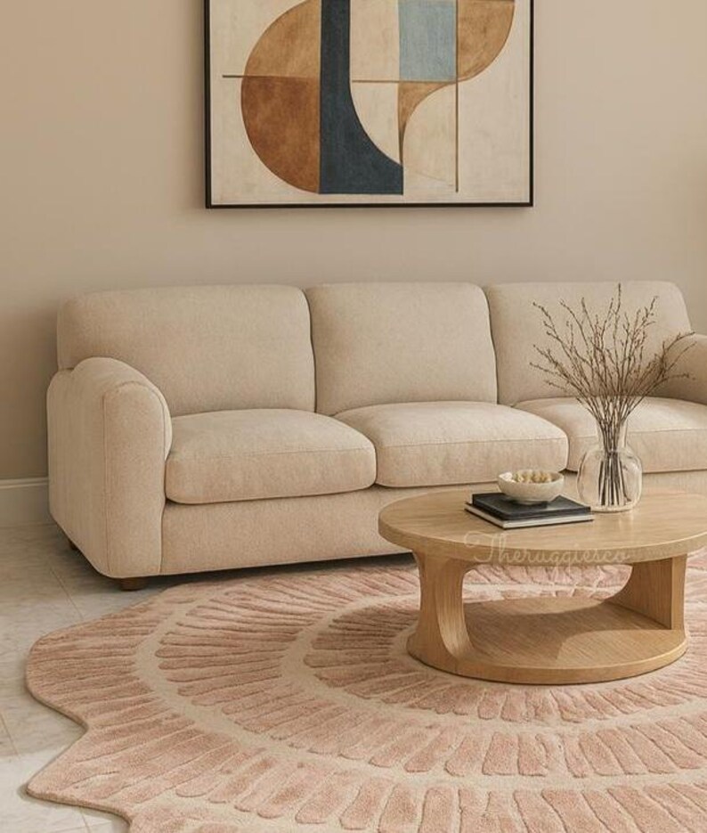 Pu&ograve; includere: Un divano beige con un tavolino rotondo in legno e un tappeto rosa. Il tappeto presenta un motivo circolare. Un vaso con rami secchi &egrave; appoggiato sul tavolo. Un dipinto astratto &egrave; appeso al muro.