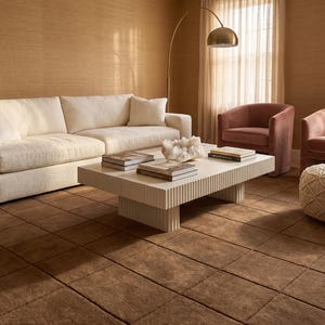 May include: A living room scene featuring a cream-colored sofa, two pink armchairs, and a textured ottoman. A white coffee table with books and decorative items sits on a brown rug. The room has a warm, inviting atmosphere.