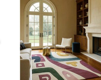 May include: A colorful area rug with an abstract design in a living room setting. The rug features a cream background with geometric shapes in green, purple, blue, and yellow. The room includes a sofa, chair, and a gold side table.