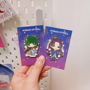 May include: Two enamel pins featuring anime-style characters, each on a purple card with the text "KOMAKI STORE". One pin has a green-haired character in a colorful outfit, the other has a purple-haired character in a blue outfit.