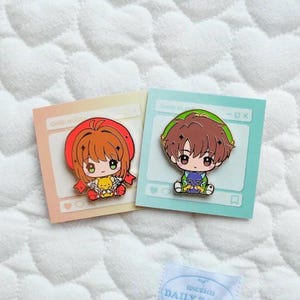 Könnte beinhalten: Zwei Emaille-Anstecknadeln mit Anime-Charakteren. Eine zeigt ein Mädchen in roter Kleidung mit einem gelben Bären, die andere einen Jungen in Grün und Braun mit einer lila Katze. Jede Nadel befindet sich auf einer pastellfarbenen Karte mit dem Text "CHIBI HUGS".