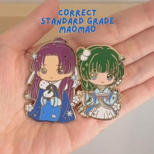 May include: Two enamel pins depicting anime-style characters. One pin shows a purple-haired character in a blue outfit, and the other a green-haired character in a blue and white outfit. The text "CORRECT STANDARD GRADE MAOMAO" is above the pins.