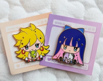 May include: Two enamel pins featuring anime characters. The first pin is yellow with a pink and white dress and a green creature. The second pin is blue with a pink and white dress and a white and purple creature. Both pins have the text "CHIBI HUGSI" on the background.