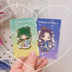 May include: Two enamel pins featuring anime-style characters. One pin has a green-haired character in a colorful kimono, the other has a purple-haired character in a blue kimono. Both pins are on cards that say "KOMAKI STORE".