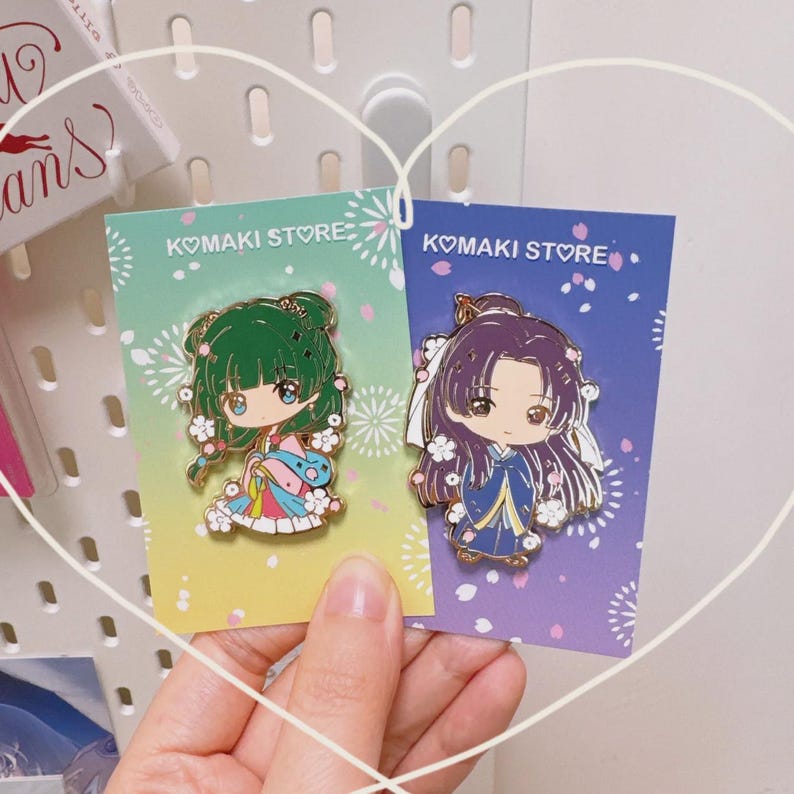 May include: Two enamel pins featuring anime-style characters. One pin has a green-haired character in a colorful kimono, the other has a purple-haired character in a blue kimono. Both pins are on cards that say "KOMAKI STORE".