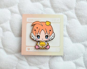 May include: A gold enamel pin featuring a cartoon character with orange hair, wearing a crown and holding a yellow chick. The pin is on a white background with a pink and orange border. The text "CHIBI MUGI" is visible at the top of the pin.