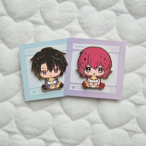 May include: Two enamel pins featuring anime-style characters with large eyes and colorful hair. One pin has a black-haired character, the other a pink-haired character. Each character is holding a small animal. The pins are on light blue and lavender cards.