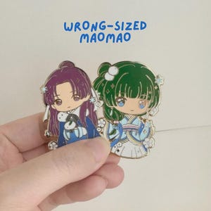 May include: Two enamel pins with anime-style characters in traditional dress. One pin features a purple-haired character, the other a green-haired character. The pins have gold borders and the text "WRONG-SIZED MAOMAO" in blue.