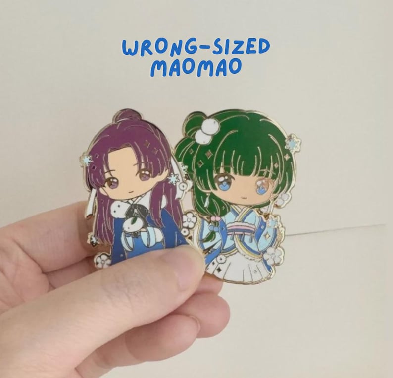 May include: Two enamel pins with anime-style characters in traditional dress. One pin features a purple-haired character, the other a green-haired character. The pins have gold borders and the text "WRONG-SIZED MAOMAO" in blue.