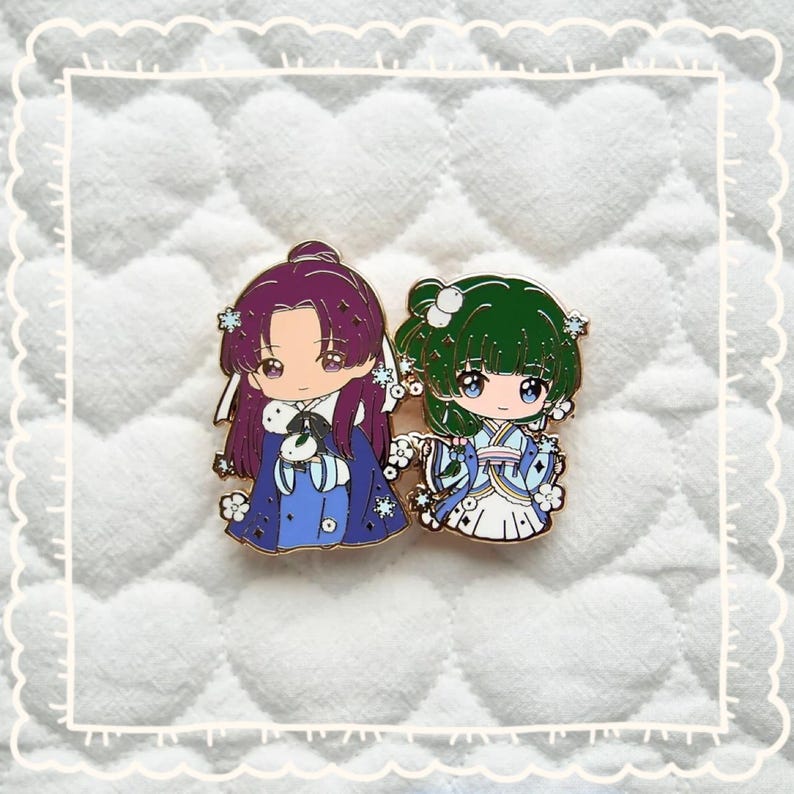 May include: Two enamel pins depicting anime-style characters. One pin shows a purple-haired character in a blue outfit, and the other has a green-haired character in a blue and white outfit. Both pins have a gold outline and are set against a white background.