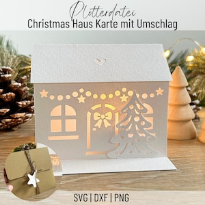 May include: A white paper Christmas card shaped like a house, with cut-out windows, a door, and a Christmas tree. The card has a heart-shaped cutout on the roof and is lit from within. The text "Christmas Haus Karte mit Umschlag" is at the top.