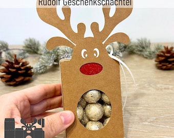 May include: A brown cardboard reindeer gift box with a cut-out window and a red nose. The box is decorated with a white ribbon and has the text "Plotterdatei Rudolf Geschenkschachtel" on the top. The box is being held by a hand and is filled with small, gold-colored balls.