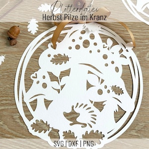 Könnte beinhalten: Weiße Papier-Ausschnitt-Dekoration mit einem kreisförmigen Design mit Herbstblättern, Pilzen und einem Igel. Der Text "Herbst Pilze im Kranz" ist sichtbar. Ein braunes Band ist zum Aufhängen befestigt. Der Text "SVG | DXF | PNG" ist sichtbar.