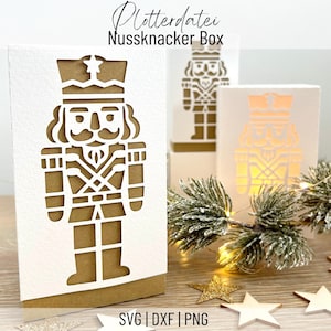 以下が含まれることがあります： 白とベージュの紙箱で、くるみ割り人形の兵士の切り抜きデザインが施されています。箱は星で飾られており、上部には「Plotterdatei Nussknacker Box」という文字があります。画像の下部には「SVG | DXF | PNG」という文字があります。