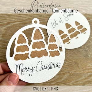 Może przedstawiać: Biała, okrągła ozdoba choinkowa z wyciętym wzorem trzech brązowych choinek i napisem "Merry Christmas". Ozdoba ma otwór na górze do zawieszenia. Na drugiej ozdobie w tle widoczny jest napis "Let it Snow".