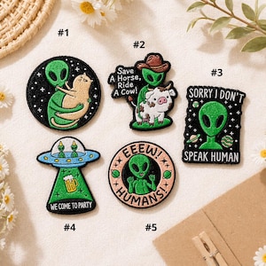 May include: A collection of embroidered patches featuring alien-themed designs. One patch shows an alien hugging a cat, another has an alien riding a cow, and a third says "Sorry I Don't Speak Human." Other patches include the phrases "We Come to Party" and "Eeeew! Humans!"