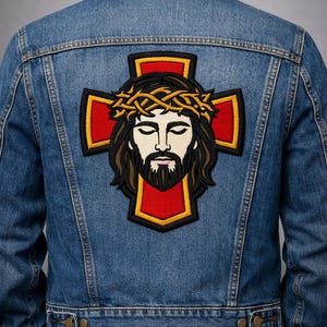 May include: A blue denim jacket with an embroidered patch on the back. The patch shows Jesus wearing a crown of thorns, set against a red cross with a yellow border. The jacket has a classic design, typical of denim jackets.