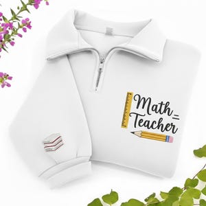May include: White pullover sweatshirt with a half-zip collar. The design features the text "Math Teacher" with a ruler and pencil graphic. The left sleeve has an embroidered stack of books.