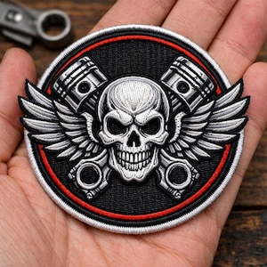 May include: Embroidered patch featuring a skull with wings and crossed pistons. The design is in shades of black, white, and gray, with a red border. The patch is round and has a biker or motorcycle theme.
