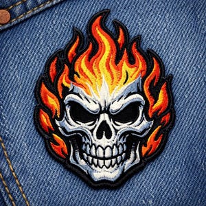 Peut inclure: Écusson brodé représentant une tête de mort en flammes. Le crâne est blanc avec des orbites noires et une mâchoire détaillée. Des flammes orange, jaune et rouge entourent le crâne, soulignées de noir. L'écusson est posé sur un fond en denim bleu.