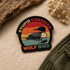 May include: Embroidered patch featuring a loon swimming on a lake, set against a sunset gradient of red, orange, yellow, and teal. The patch reads "STUPID CANADIAN WOLF BIRD" in a circular design.