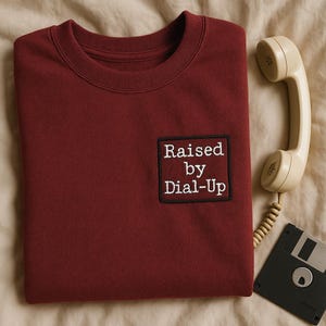 May include: A maroon sweatshirt with the text "Raised by Dial-Up" embroidered on a patch. A vintage beige telephone handset and a floppy disk are next to the sweatshirt on a beige surface.