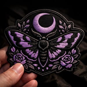 May include: Embroidered patch featuring a moth with purple and black wings, a crescent moon, and roses. The patch has a black background and a decorative border. The moth's wings have a detailed pattern.