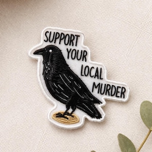 May include: Embroidered patch featuring a black raven with the text "SUPPORT YOUR LOCAL MURDER" in black letters on a white background. The raven is perched on a gold-colored base, with a white border.