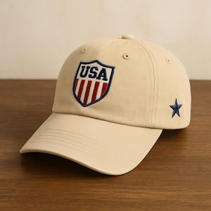May include: A tan baseball cap with a curved brim. The cap features a shield-shaped emblem with the word "USA" and red and white stripes. A navy blue star is embroidered on the side. The cap is made of a soft material.