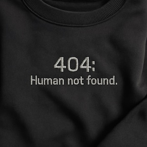 May include: Black sweatshirt featuring the embroidered phrase "404: Human not found." The text is in white, sans-serif lettering. The sweatshirt has a ribbed crew neck and a soft, textured appearance.