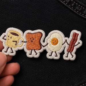 May include: Embroidered patch featuring a coffee cup, toast, fried egg, and bacon, each with smiling faces and holding hands. The patch has a white border and is set against a black background.