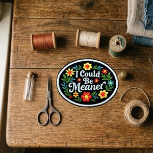May include: A black oval patch embroidered with the words "I Could Be Meaner" and floral designs. The image shows sewing tools and materials, such as thread spools, scissors, and a thimble, on a wooden table.