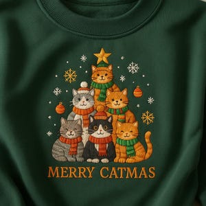 May include: A dark green sweatshirt featuring an embroidered design of cats arranged in the shape of a Christmas tree. The cats wear scarves and hats, with snowflakes and a star. The text "MERRY CATMAS" is embroidered below the cats.