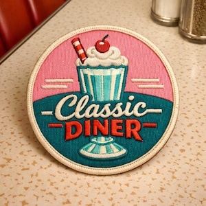 May include: Embroidered patch with a retro diner theme. The design features a milkshake with a cherry on top, and the words "Classic Diner" in red and white. The patch has a pink and teal colour scheme.