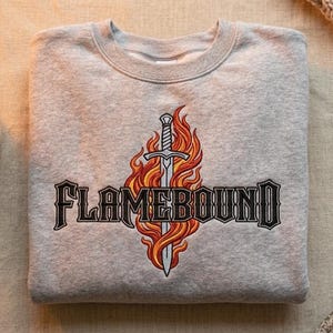 May include: A light gray sweatshirt with the word "FLAMEBOUND" in a black, gothic font. A sword is centered above the text, surrounded by orange and red flames. The sweatshirt is folded neatly on a neutral surface.