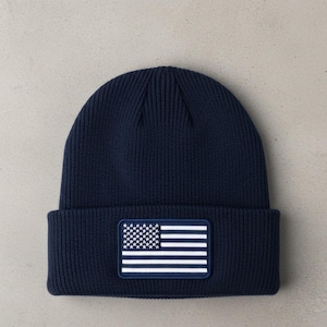 Peut inclure: Bonnet en tricot bleu marine avec un écusson rectangulaire représentant le drapeau américain. Le bonnet a une texture côtelée et un bord replié. L'écusson du drapeau affiche des étoiles et des rayures blanches sur fond bleu marine.