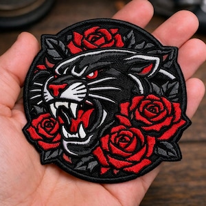 May include: Embroidered patch featuring a black panther with red eyes and open mouth, surrounded by red roses and green leaves. The patch has a black border and is likely for clothing or accessories.