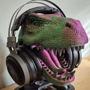 May include: A green and pink dinosaur head headset stand with a black gaming controller and headphones.