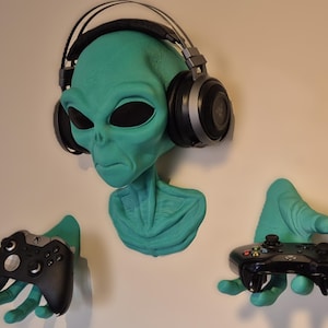 May include: A teal alien head wall mount with black headphones. Two teal alien hands hold black gaming controllers. The alien head has large black eyes and is mounted on a white wall. A unique gaming accessory.